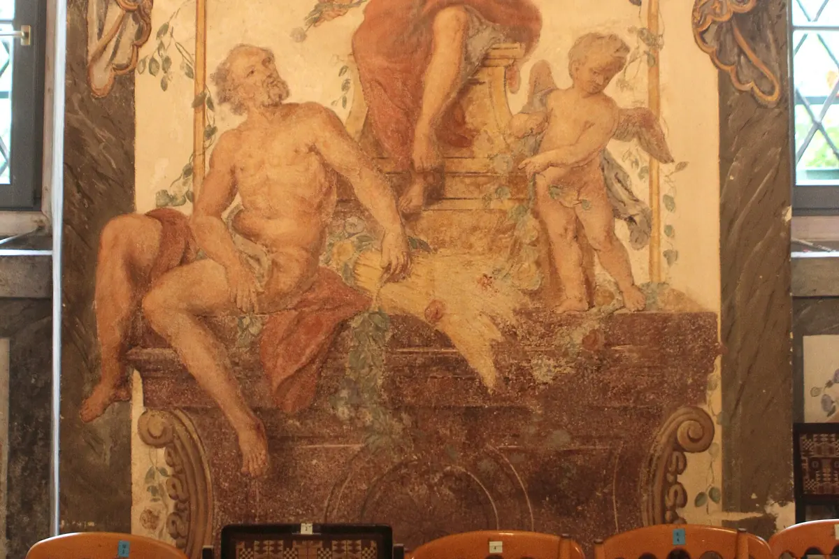 A painted wall panel showing a reclining elderly man and a winged cherub beside a sheaf of wheat, with leaded windows on either side and the backs of concert chairs below.